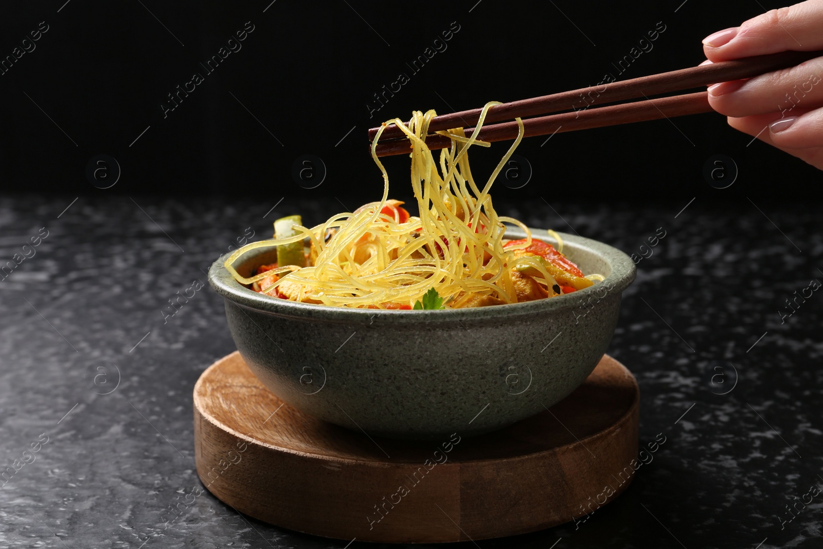 Photo of Stir-fry. Woman using chopsticks for eating tasty noodles with meat at dark textured table, closeup