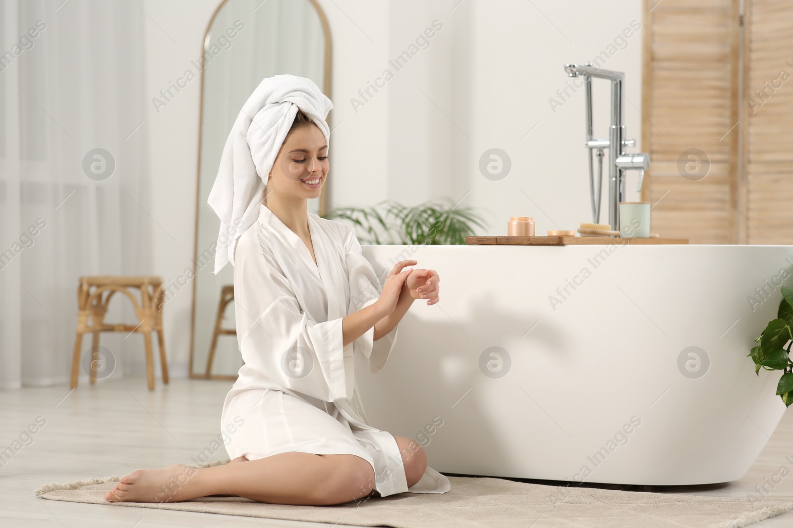 Photo of Beautiful happy woman in white robe applying cream near tub in bathroom