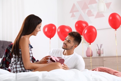 Photo of Young man presenting gift to his girlfriend in bedroom decorated with air balloons. Celebration of Saint Valentine's Day