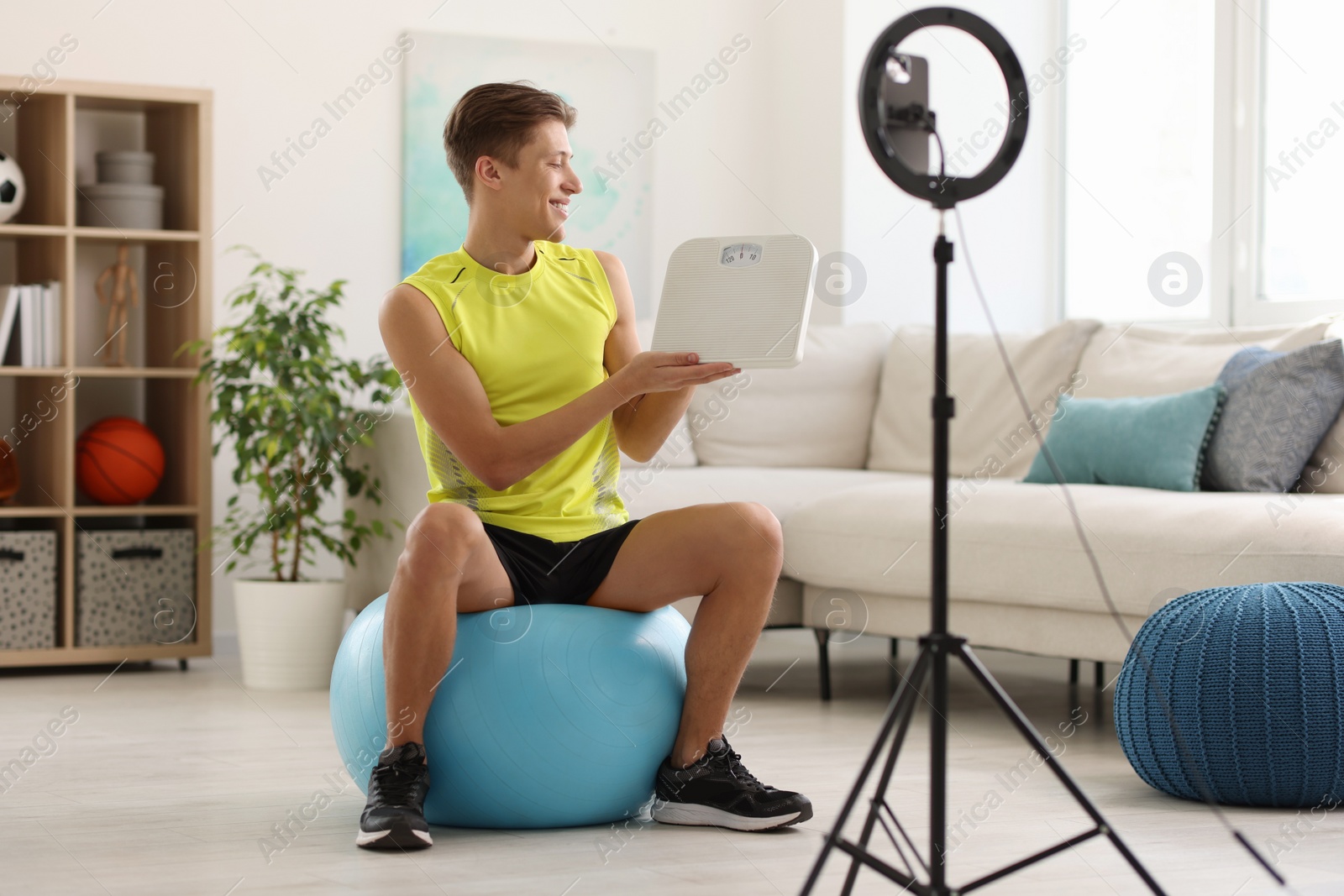 Photo of Smiling sports blogger holding floor scales while streaming online fitness lesson with phone at home
