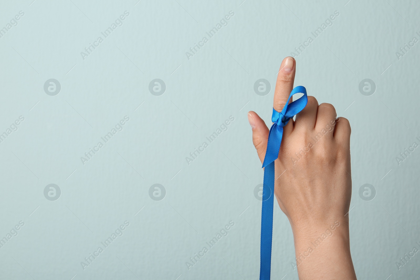 Photo of Woman with blue ribbon on finger against light background, closeup. Symbol of social and medical issues