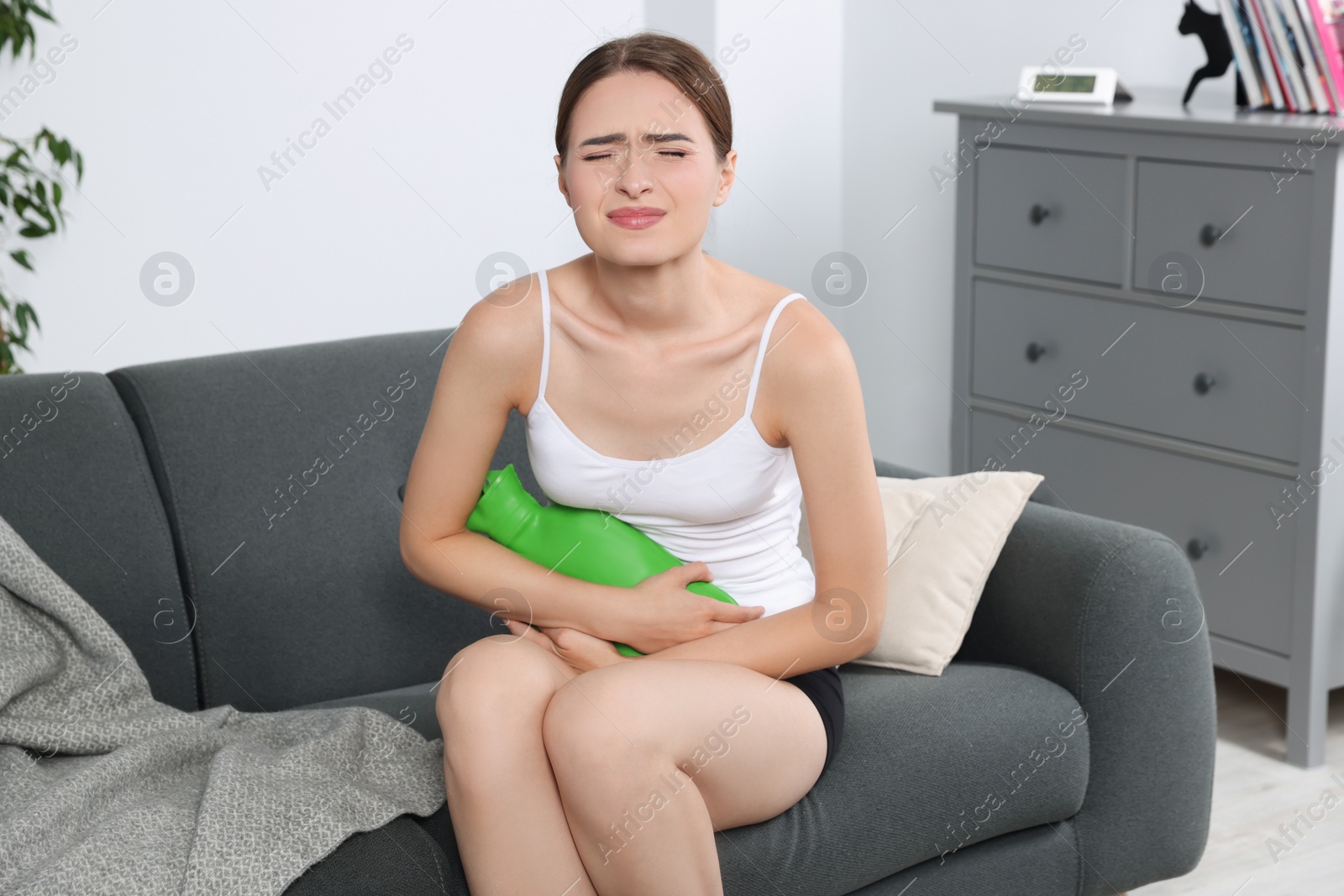 Photo of Young woman using hot water bottle to relieve cystitis pain on sofa at home