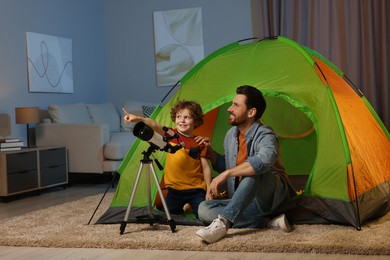 Photo of Father and his son using telescope to look at stars while sitting in camping tent indoors