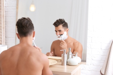 Photo of Young man shaving near mirror in bathroom