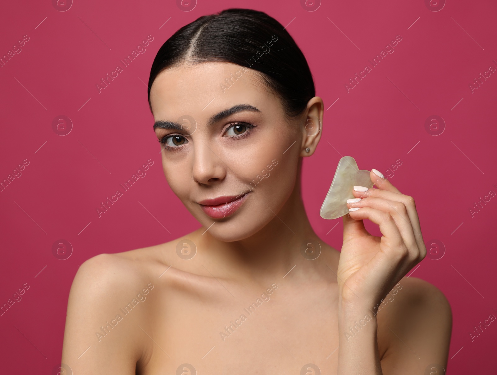 Photo of Beautiful young woman doing facial massage with gua sha tool on pink background