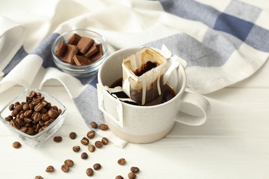 Photo of Cup with drip coffee bag and beans on white wooden table, closeup