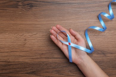 Woman holding light blue ribbon with paper blood drop at wooden table, top view and space for text. Diabetes awareness