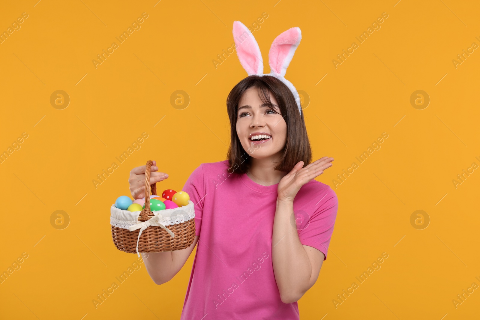 Photo of Easter celebration. Happy woman with bunny ears and wicker basket full of painted eggs on orange background