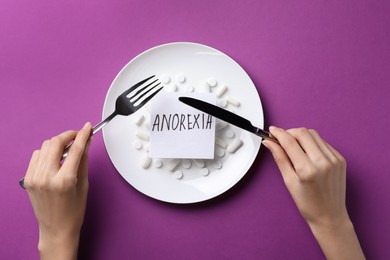 Photo of Woman holding cutlery over plate with pills and word Anorexia on purple background, top view