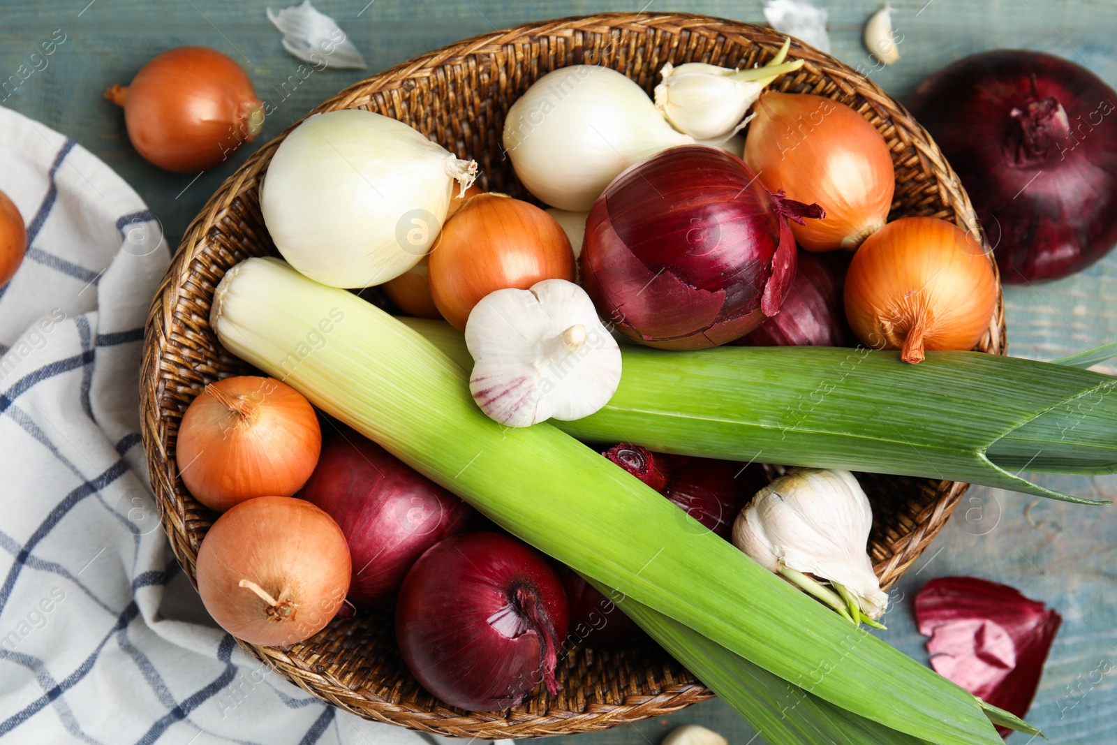 Photo of Wicker basket with fresh onion bulbs, leeks and garlic on light blue wooden table, flat lay