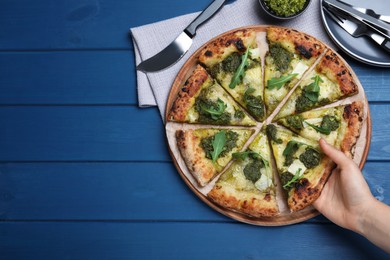 Photo of Woman taking slice of delicious pizza with pesto, cheese and arugula at blue wooden table, closeup. Space for text