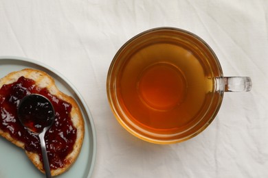 Photo of Cup of freshly brewed tea and bread with jam on white fabric, flat lay