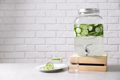 Photo of Jar dispenser of fresh cucumber water and glasses on table against brick wall. Space for text
