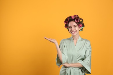 Photo of Happy young woman in silk bathrobe with hair curlers on yellow background. Space for text