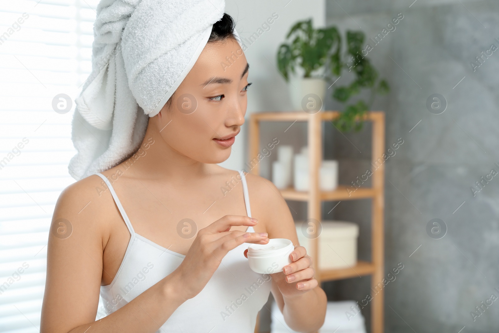 Photo of Beautiful young Asian woman holding body cream in room, space for text