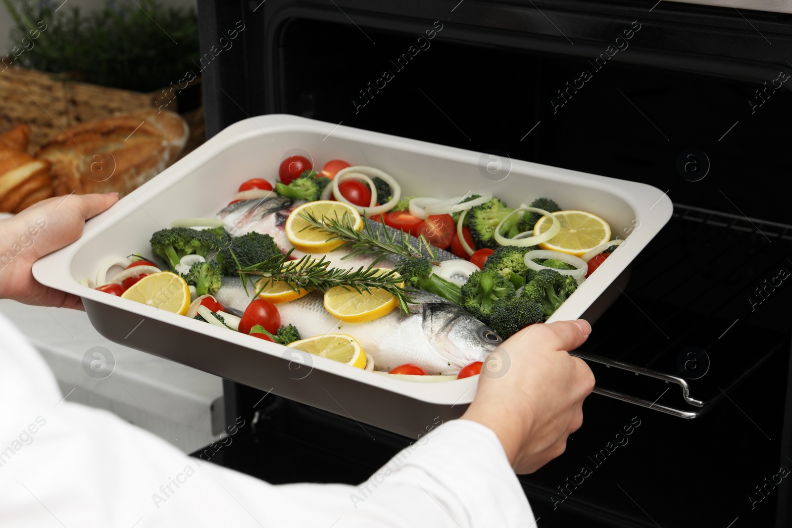 Photo of Woman putting baking dish with raw fish and vegetables into oven in kitchen, closeup