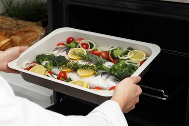 Photo of Woman putting baking dish with raw fish and vegetables into oven in kitchen, closeup