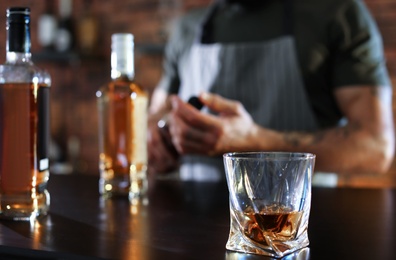 Bartender at counter with bottles and glass of whiskey in bar, closeup. Space for text