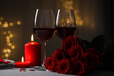 Glasses of red wine, burning candles and rose flowers on grey table against blurred lights. Romantic atmosphere