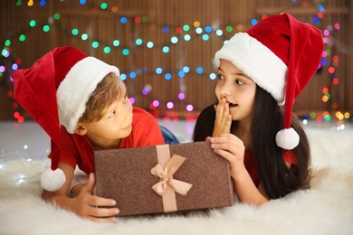 Photo of Cute little children in Santa hats opening Christmas gift box on floor