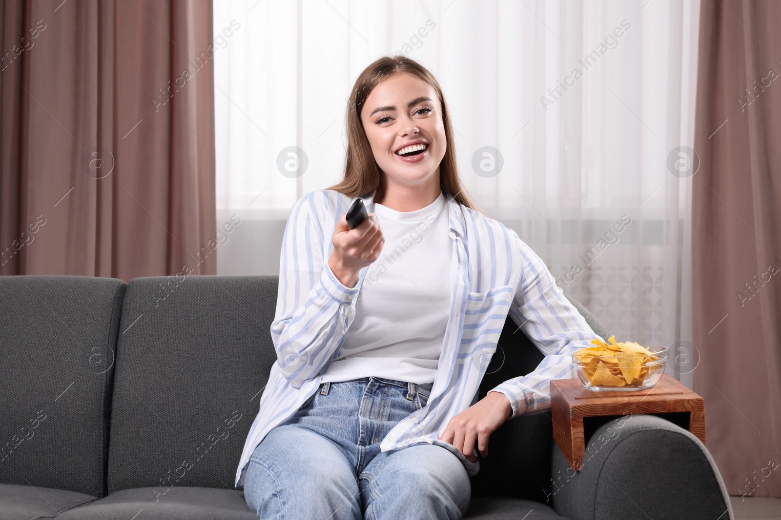 Photo of Happy woman switching TV channels at home. Nacho chips on wooden sofa armrest table