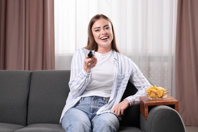 Happy woman switching TV channels at home. Nacho chips on wooden sofa armrest table