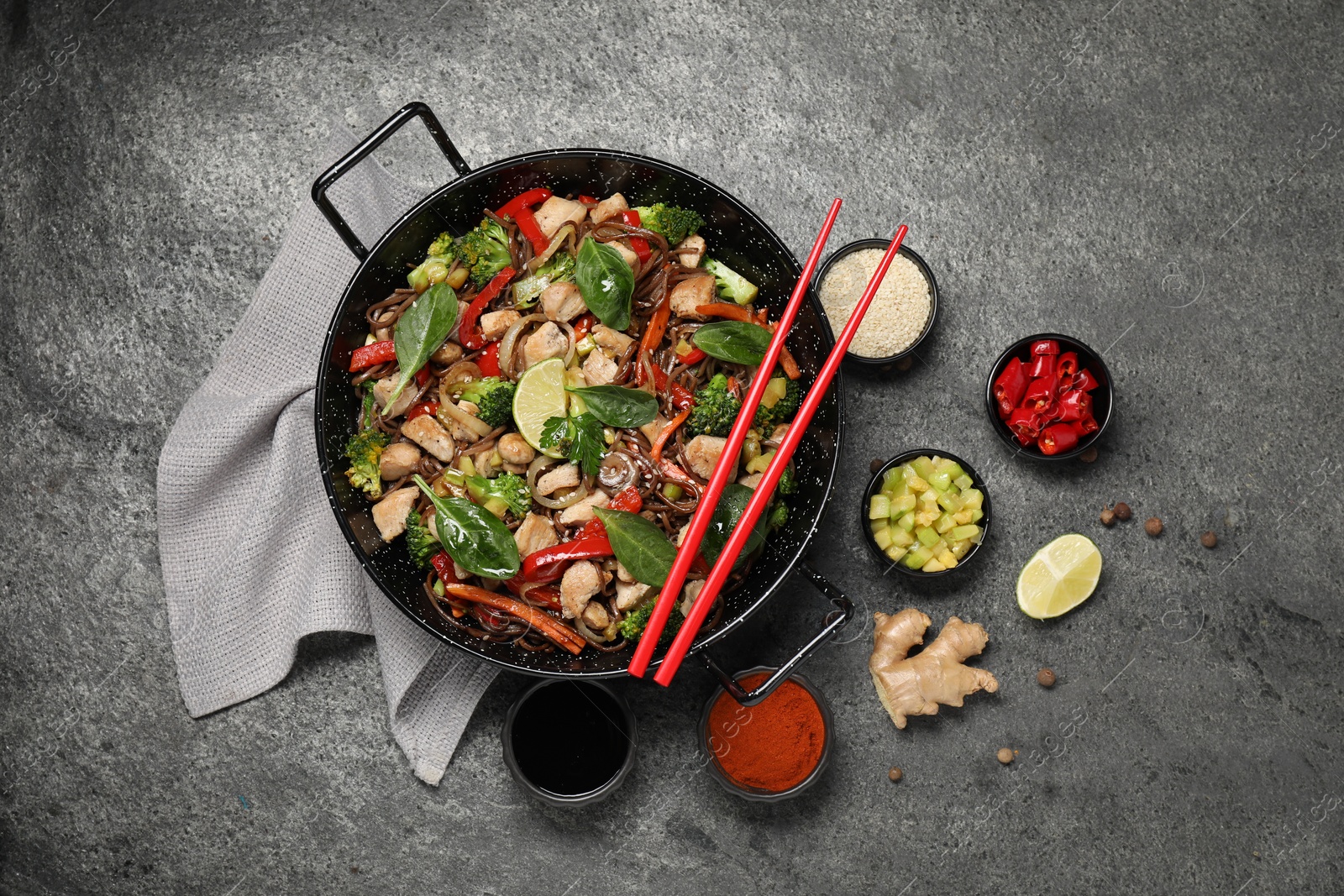 Photo of Stir-fry. Tasty noodles with meat in wok, chopsticks and ingredients on grey textured table, flat lay