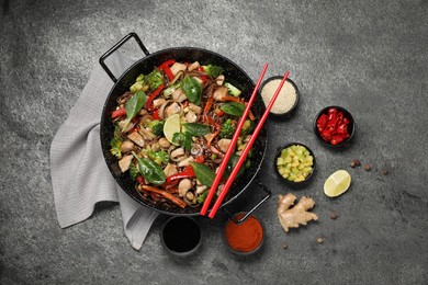 Stir-fry. Tasty noodles with meat in wok, chopsticks and ingredients on grey textured table, flat lay