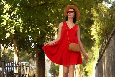 Beautiful young woman in stylish red dress and sunglasses walking on city street