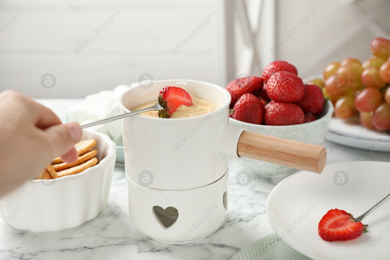 Photo of Woman dipping strawberry into fondue pot with white chocolate at marble table