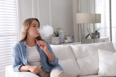 Photo of Beautiful young woman using disposable electronic cigarette on sofa at home