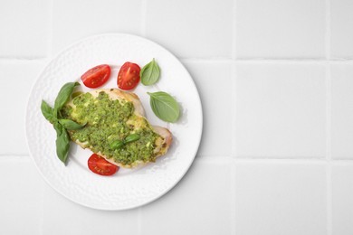 Photo of Delicious chicken breast with pesto sauce, tomatoes and basil on white tiled table, top view. Space for text