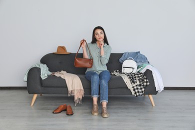 Photo of Thoughtful young woman holding bag on sofa with clothes indoors