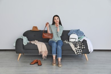 Thoughtful young woman holding bag on sofa with clothes indoors