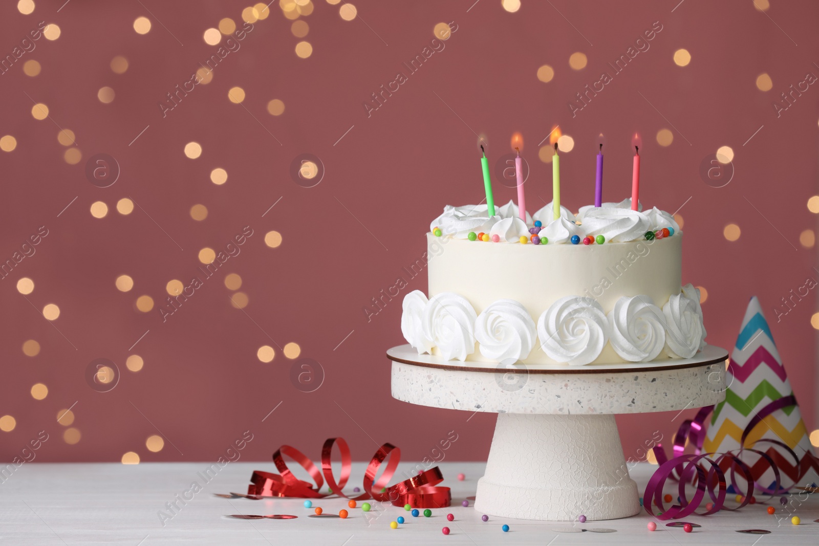Photo of Birthday cake with burning candles and decor on white table against blurred festive lights, space for text