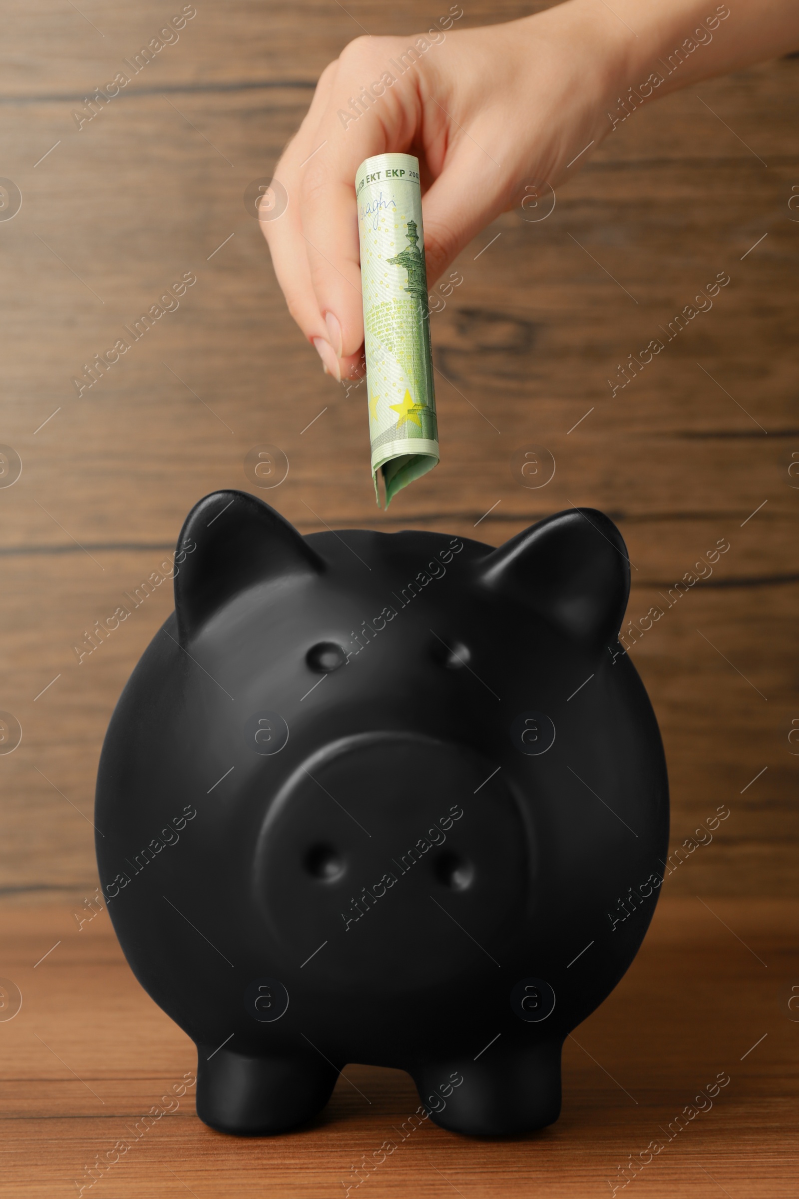 Photo of Woman putting banknotes into piggy bank at wooden table, closeup