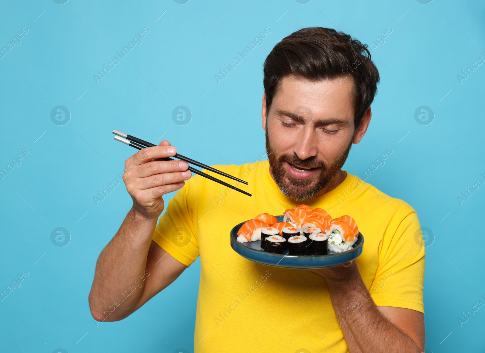 Photo of Happy man holding plate with tasty sushi rolls and chopsticks on light blue background