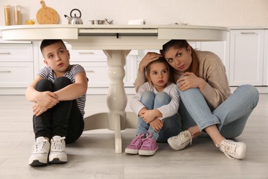 Photo of Scared mother with her children hiding under table in kitchen during earthquake