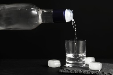 Photo of Pouring vodka from bottle in glass and ice cubes on black table