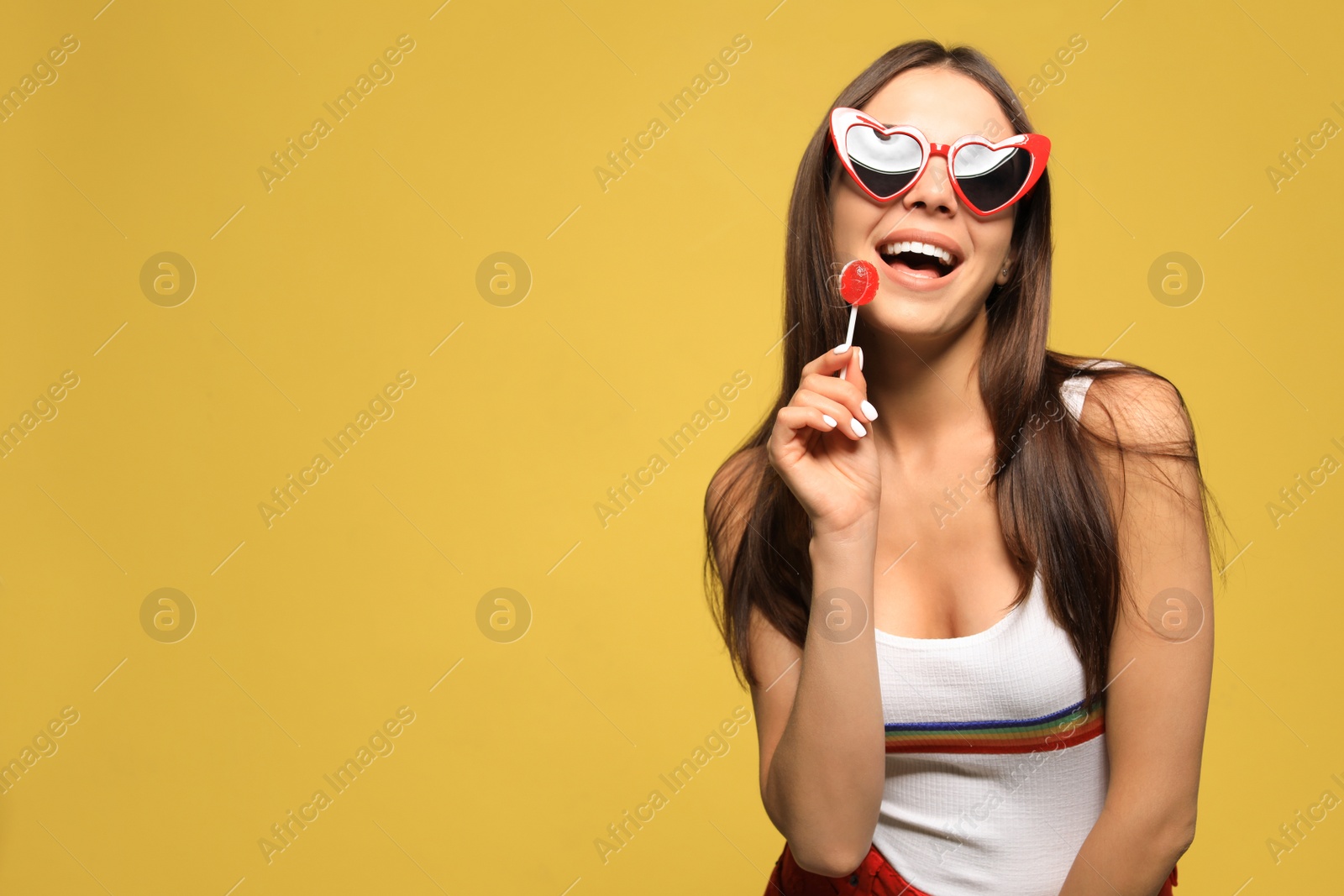 Photo of Portrait of beautiful young woman with heart shaped sunglasses and lollipop on color background. Space for text