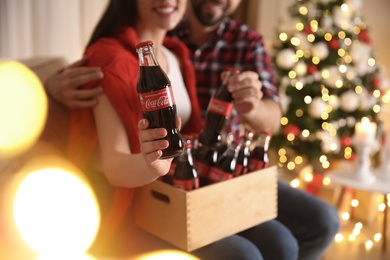MYKOLAIV, UKRAINE - JANUARY 27, 2021: Young couple holding crate with bottles of Coca-Cola in room decorated for Christmas, closeup