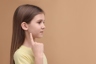 Photo of Hearing problem. Little girl pointing at ear on pale brown background, space for text