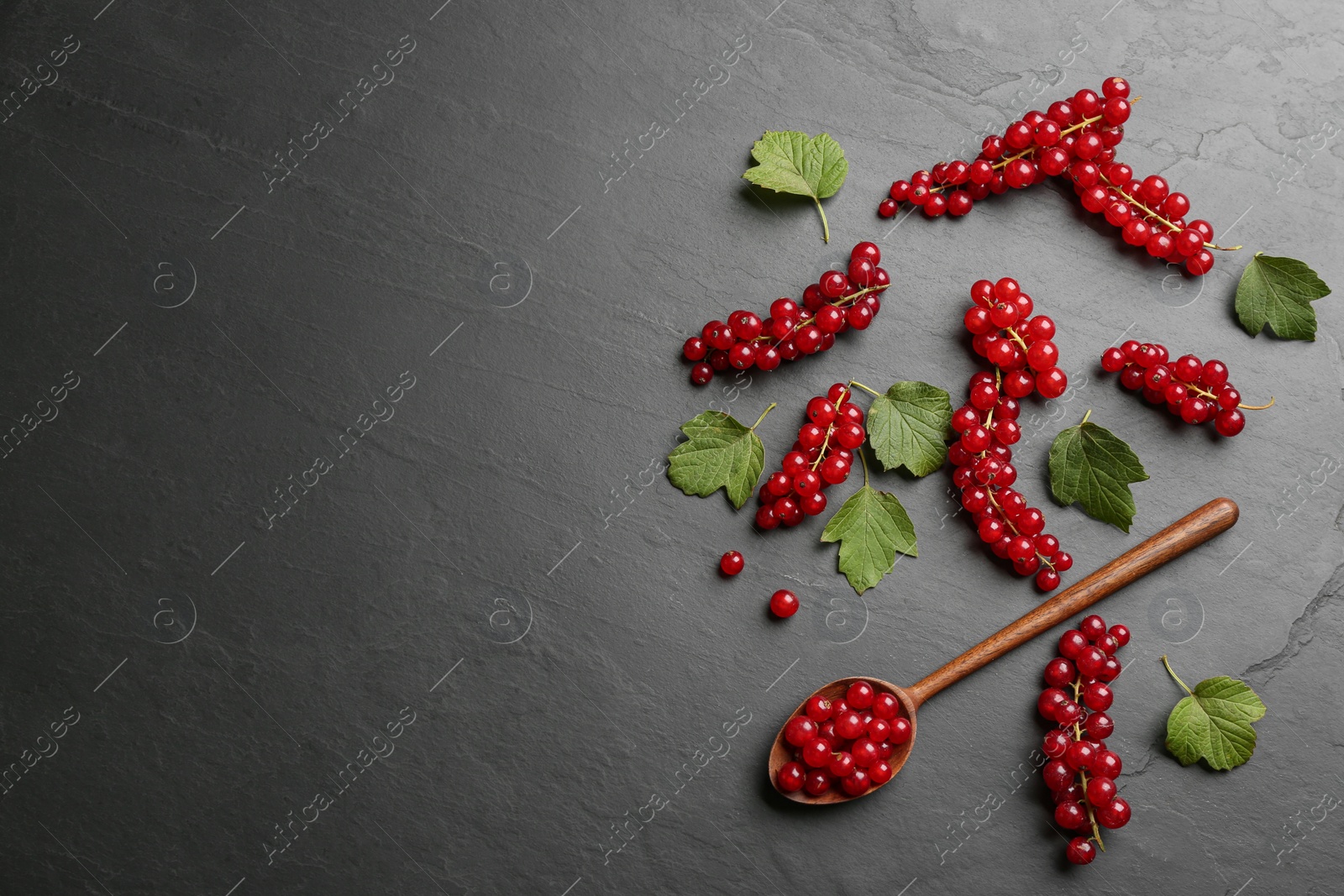 Photo of Delicious red currants, leaves and wooden spoon on black slate table, flat lay. Space for text