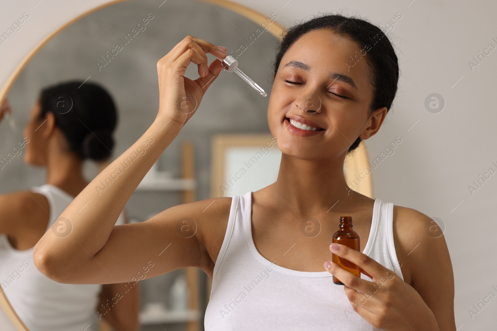 Photo of Smiling woman applying serum onto her face in bathroom