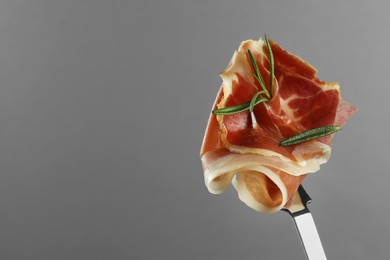 Photo of Fork with slice of tasty jamon and rosemary on light grey background, closeup. Space for text