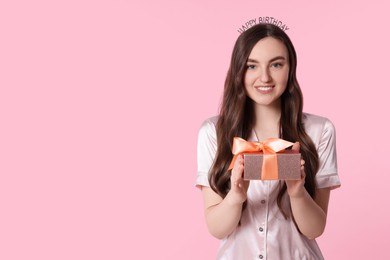 Beautiful young woman in headband holding gift box on pink background, space for text. Happy Birthday
