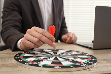 Photo of Business targeting concept. Man with dart aiming at dartboard at wooden table indoors, closeup