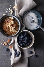 Photo of Tasty granola in bowl, blueberries, yogurt and spoon on gray textured table, flat lay