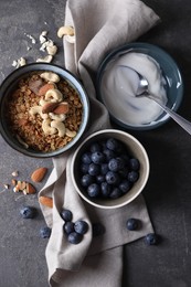 Tasty granola in bowl, blueberries, yogurt and spoon on gray textured table, flat lay