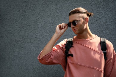 Photo of Handsome young man in stylish sunglasses and backpack near grey wall, space for text